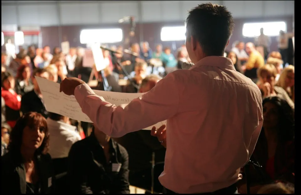 Facilitator holding up a sheet of paper in front of a large seated audience